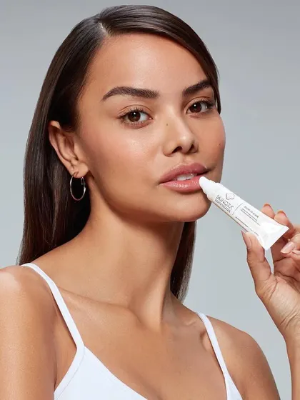 A focused application shot for a lip treatment product. The model is shown applying the balm from a tube, highlighting the product's ease of use and its direct benefit for lip care.