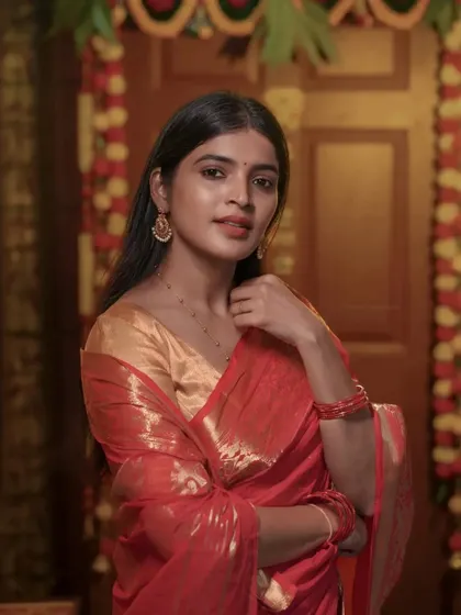 A close-up portrait focusing on the actress's expression. The warm lighting complements the red saree and gold blouse perfectly.