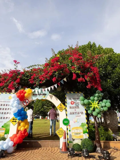 The entrance to a "Wheels on the Bus" themed party, set against a beautiful backdrop of green trees and bright red bougainvillea. The colorful balloon arch and themed signs create a cheerful welcome.
