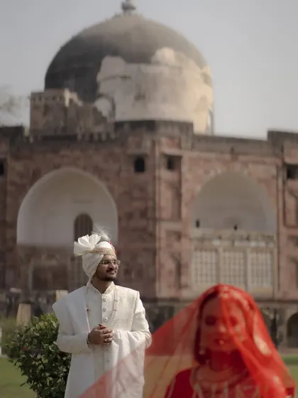 A dramatic and artistic portrait. The groom stands in sharp focus while the bride is a vibrant, soft-focus figure in red in the foreground.