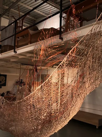 A large, hammock-like sculpture woven from copper and natural fibers, suspended in our studio for the 'Cu+Fibers' exhibition. Its form is a result of a continuous dialogue between intent and material.