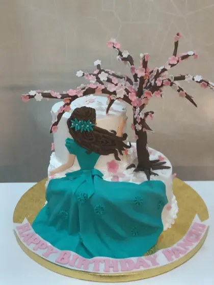 A serene and artistic cake featuring a fondant figure of a woman sitting under a delicate, handcrafted cherry blossom tree.