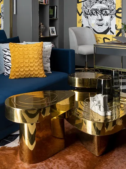 A close-up of a bold and eclectic living space. A royal blue sofa is paired with a vibrant yellow cushion and a sculptural, high-shine gold coffee table, showcasing our ability to create customized, personality-driven furniture and spaces.