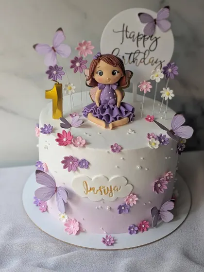 A fairy-themed cake for a first birthday. This pretty design in shades of purple features a cute fairy figurine, delicate flowers, and fluttering butterflies.