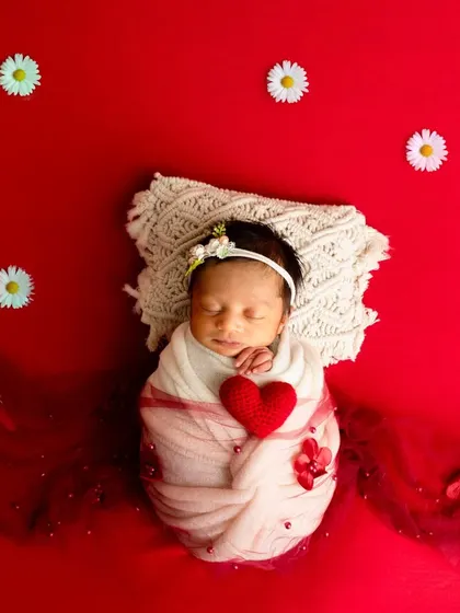 A newborn swaddled in white holds a red heart, lying on a red background dotted with white daisies for a classic, high-contrast look.