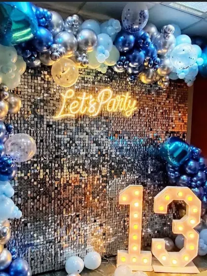 A 13th birthday party setup with a silver shimmer wall and a blue and silver balloon arch. Marquee numbers indicate the milestone age.