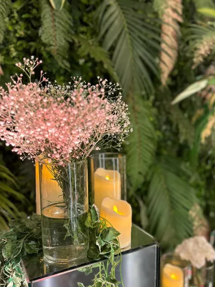 A simple yet elegant detail: a vase of pink baby's breath and pillar candles on a mirrored cube, set against a wall of green leaves.