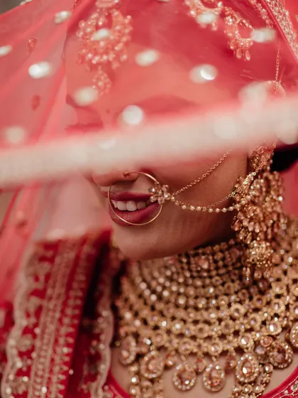 A close-up shot of the bride's smile, partially hidden by her red veil, capturing her happiness and the intricate details of her nath and necklace.