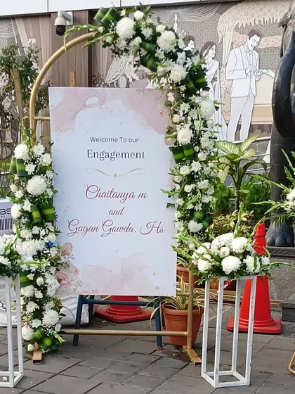 A modern welcome sign for an engagement, framed by a golden arch and white floral arrangements.