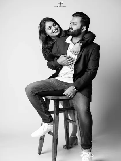 A classic black and white portrait of a couple on a stool. The woman's embrace from behind and their shared look create a feeling of warmth and connection.