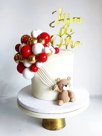 A simple and sweet teddy bear cake for Jija's first birthday. The red, white, and gold balloon cascade adds a festive touch to the clean white design.