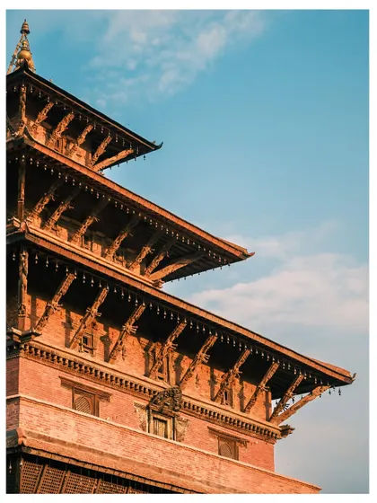 The detailed, multi-tiered roof of a traditional Newari pagoda in Kathmandu. The warm sunlight highlights the intricate wooden carvings against a brilliant blue sky.