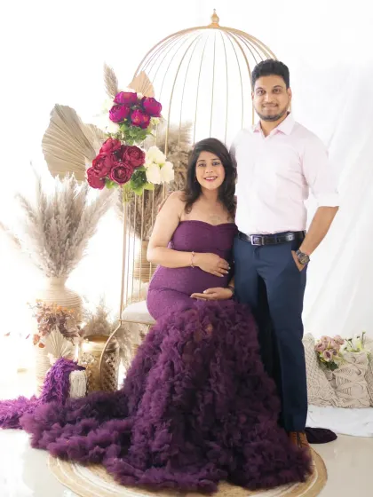 An elegant couple's portrait in our boho floral setup. The dad-to-be stands proudly beside his partner, who is seated and wearing a beautiful purple gown.