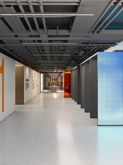 This corridor in a modern workplace features an exposed ceiling, revealing the raw, honest infrastructure of the building. This industrial aesthetic is balanced by clean white walls, bold orange graphics, and a dynamic LED screen, creating a space that is both exuberant and authentic.