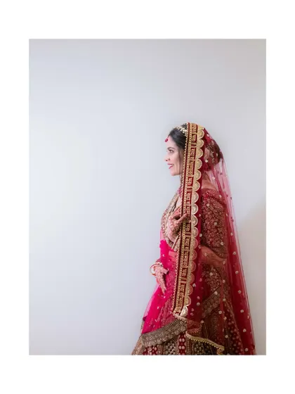 A minimalist and elegant bridal portrait. The bride stands against a plain white wall, her vibrant red lehenga and graceful profile taking center stage.