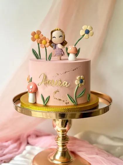 A whimsical garden cake with a girl figure, tall flowers, and mushrooms. The gold splatter on the pink buttercream adds a touch of elegance.