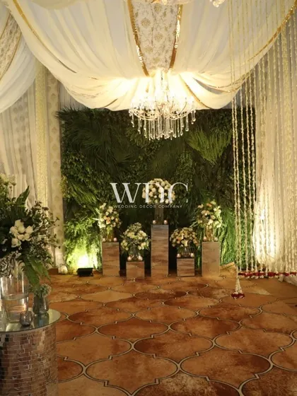 A beautiful corner featuring a wall of greenery, elegant white drapes, and cascading floral strings, all under the warm glow of a grand chandelier.