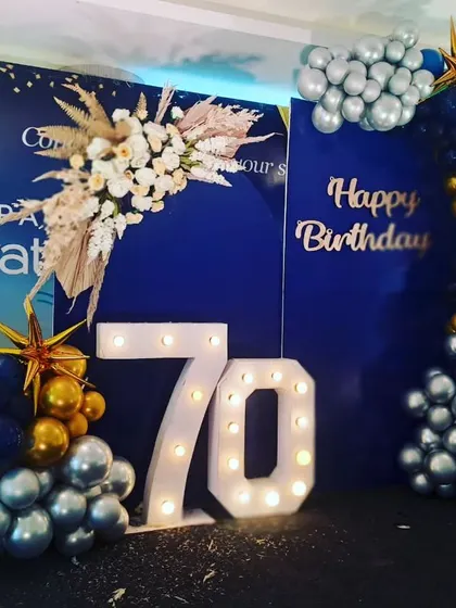 A sophisticated decor for a 70th birthday party. We used a royal blue backdrop with a gold and silver balloon garland and large light-up marquee numbers to create a celebratory and elegant look.