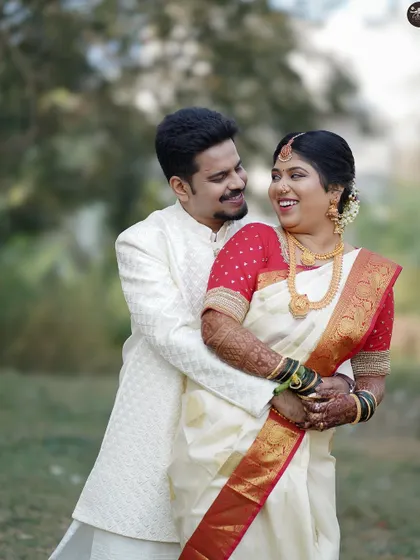 A joyful, candid shot of a couple sharing a laugh during their outdoor wedding photoshoot. The bride is wearing a traditional white and red saree, complemented by the groom's white sherwani.