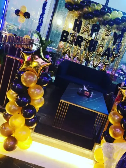 An elegant black and gold birthday setup for 'Aarvi' at a rooftop lounge, using balloon pillars topped with star foils to frame the seating area.