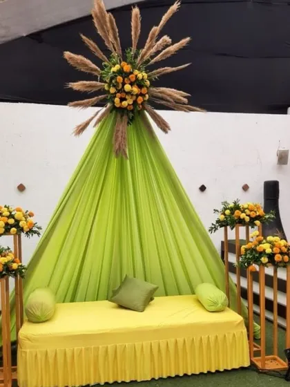 A unique and modern Haldi decoration idea for your home or terrace. This setup features a lime-green tent-style backdrop with a sunburst floral arrangement made of marigolds and pampas grass.