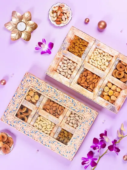 An elegant flatlay of two rustic-style dry fruit gift boxes on a pastel purple background. The scattered flowers and pearls add a delicate and premium touch to the festive packaging.
