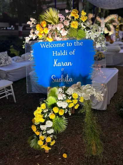 A personalized welcome sign for Karan and Suchita's Haldi, featuring a bold blue brushstroke on a clear acrylic board, framed with flowers.