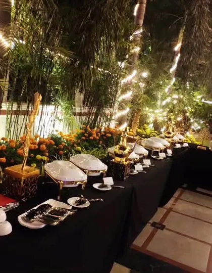 An elegant buffet arrangement for an evening event, set amidst lush greenery and fairy lights. The display features white ceramic serving dishes and ornate gold-accented warmers.