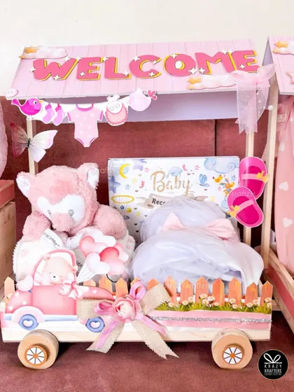 A close-up of one of the carriages from the 'Welcome Baby' train, filled with a soft teddy bear, baby record book, and other adorable pink-themed gifts.