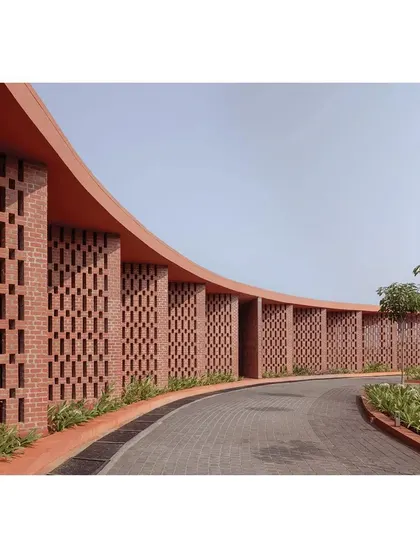 The sweeping curve of the brick jali wall guides visitors along the path to the main entrance. The intricate brickwork patterns create a dynamic play of light and shadow, a core element of this public architecture project.
