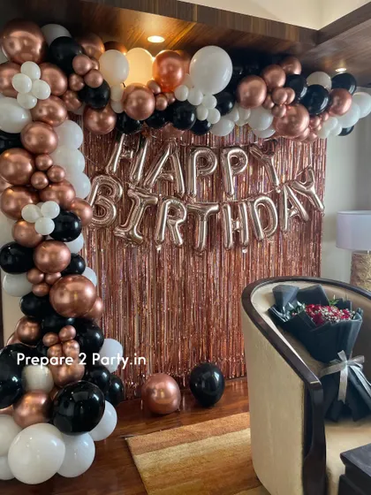 A classic and stylish birthday decoration for a home setting. This design uses a rose gold fringe curtain, a "Happy Birthday" foil balloon banner, and a balloon arch in rose gold, black, and white.
