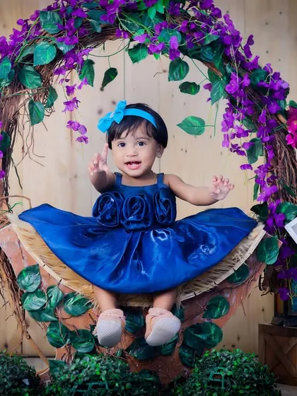 A one-year-old girl in a beautiful blue dress, sitting in a basket adorned with a wreath of purple flowers. A classic and elegant milestone portrait.