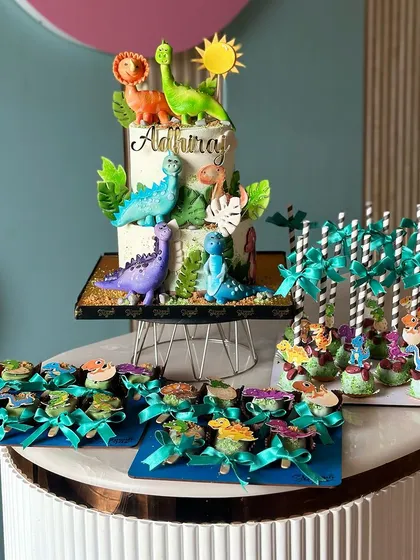 A full view of our dinosaur themed dessert table. It features a two tiered cake with adorable dino figures, along with matching cake pops and cakesicles, creating a prehistoric party scene.