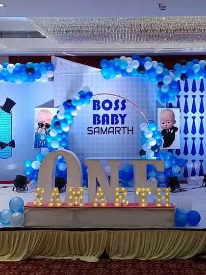A grand Boss Baby themed stage decoration perfect for a first birthday party. This large-scale setup features multiple themed backdrops, extensive blue and white balloon arches, character cutouts, and illuminated marquee letters spelling out the birthday boy's name.