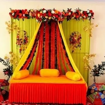 A traditional Mehendi seating arrangement with a canopy-style draping of yellow and red fabric over a comfortable diwan. The backdrop uses a fresh lime green color.