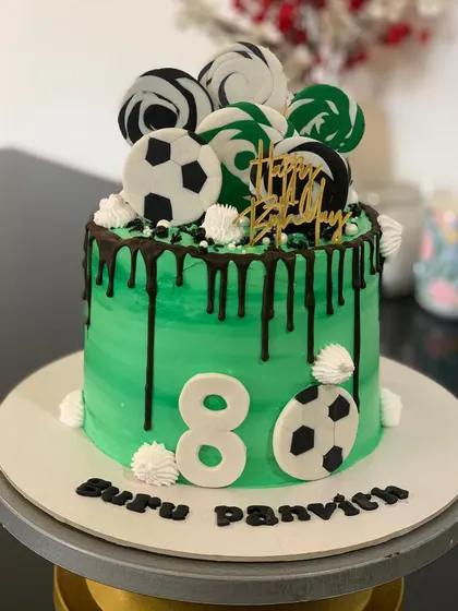 A stylish green football cake with a chocolate drip. The top is decorated with football-themed lollipops and chocolate swirls.