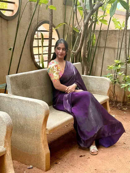 A candid moment, sitting on a bench in a garden. The soft fabric of the purple saree drapes so beautifully, making it feel both elegant and comfortable for a day out.