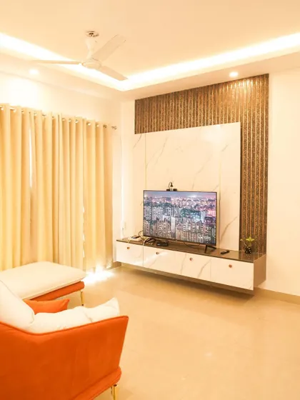 A bright and airy living room with an orange accent sofa. The TV is mounted on a wall featuring a metallic fluted panel and a white marble-finish backdrop.