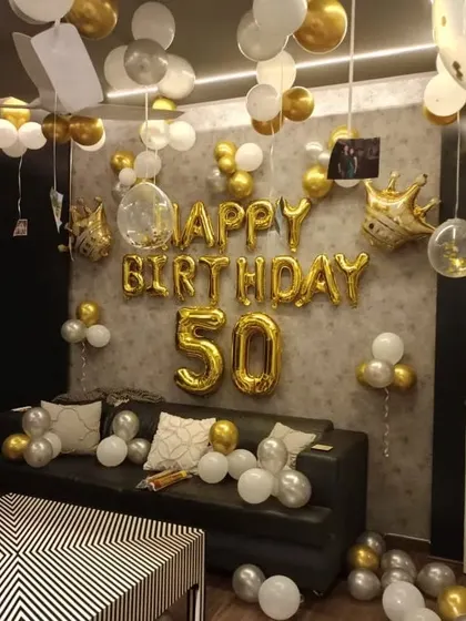 A 50th birthday celebration at home with gold and white balloons filling the room. Gold foil balloons spell out 'HAPPY BIRTHDAY 50'.