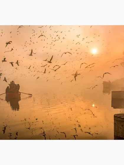 A wider perspective of the morning at Yamuna Ghat. The image balances the photographer on the right with a boat on the left, all within a beautiful, misty sunrise and a sky full of birds.