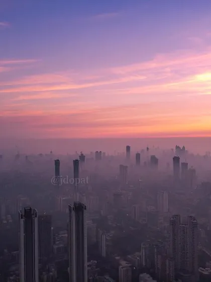 A high-altitude drone shot shows the clean, pink and purple sky above a thick layer of smog covering Mumbai.