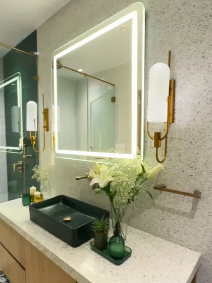 This bathroom design focuses on clean lines and a refreshing color palette. A deep green vessel sink sits atop a terrazzo-style countertop, flanked by elegant wall sconces. The large, backlit mirror provides excellent, even lighting, making the space feel bright and open.