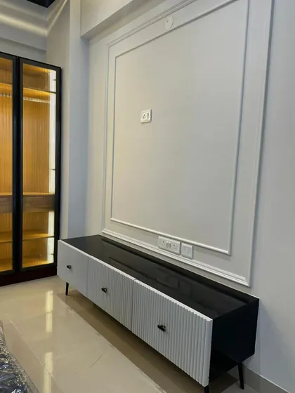 A minimalist TV console with a black top and fluted white drawers, set against a wall with classic moulding. To the side, a tall, wood-finished display cabinet with integrated lighting offers elegant storage.