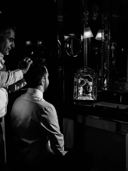 A classic, moody black and white shot of the groom getting ready. The reflections in the mirror create a complex, layered composition, capturing his anticipation in a timeless style.