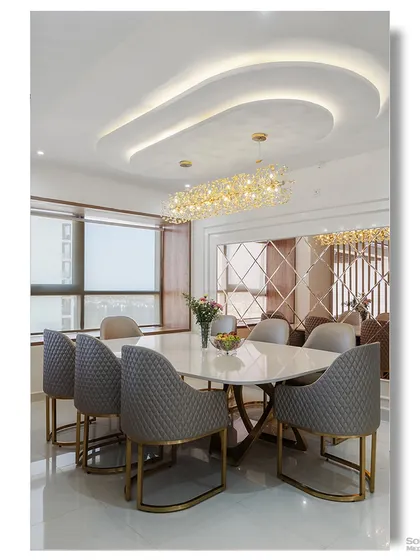 A luxurious dining room featuring quilted grey chairs, a marble table, and a stunning linear crystal chandelier. The curved ceiling detail and mirrored wall add layers of sophistication and glamour.