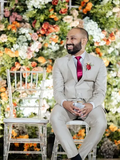 The groom enjoying the reception speeches. The backdrop of a dense floral wall adds texture and color, enhancing the celebratory atmosphere of the event.