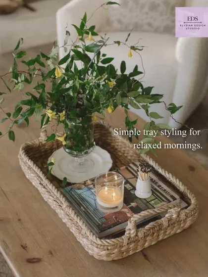 Simple tray styling for relaxed mornings. A woven tray on a coffee table holds a candle, a book, and a small plant, showing how a few curated items can create a beautiful and calming vignette.