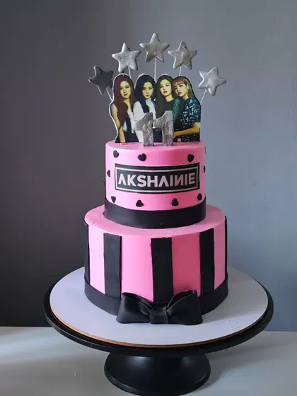 A two-tier cake for a Blackpink fan, designed in the band's signature pink and black colors. This whipped cream cake is decorated with fondant stripes, a bow, and a topper featuring the group.