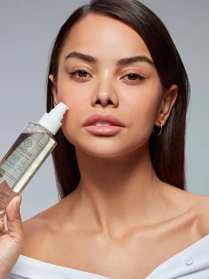 A close-up beauty shot of a model holding a clear facial toner or mist. The focus is on the product's purity and the model's hydrated, glowing skin, communicating a sense of freshness.