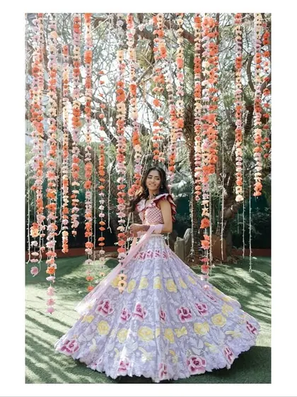 A bride stands under a curtain of flowers at her carnival-themed event. The vibrant colors and her beautiful lehenga create a whimsical and joyful image.